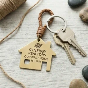 A detailed, square-format photograph focusing on a house-shaped brass custom engraved keychains, engraved with realtor branding and our first home text, resting next to a set of house keys.